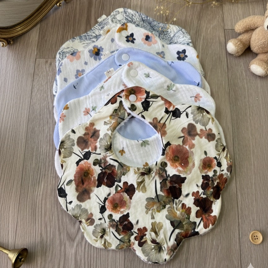 Coffret de 5 bavoirs bébé forme fleur en gaze de coton à motifs floraux et pastels, posés sur parquet en bois clair.